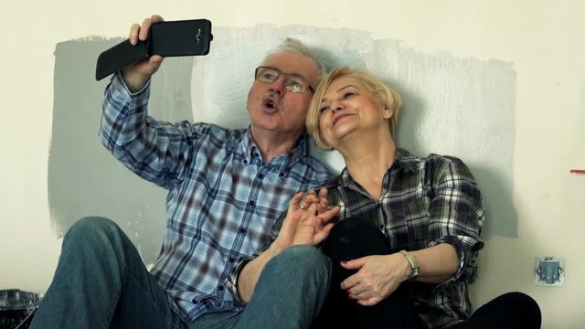 Mature couple taking selfie photo with cellphone sitting on floor at their home
