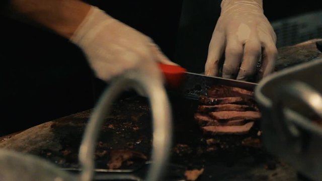 Close up on hands holding a smoked brisket on a cutting board and quickly slicing cooked beef meat into thin slices for a barbecue plate in a restaurant.
