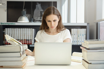 Portrait of female student sitting at the bookshelf and using laptop in the library, reading books,...