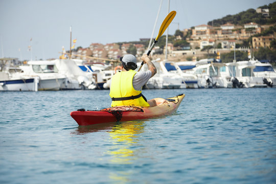Fresh and amazing picture of kayaking young man in yellow safety vest. Gorgeous landscape of seaside view, yachts and houses on the rise. Lonely man on canoe paddling on the blue sea in sunny day.
