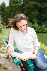 Obraz premium Young woman sitting near pond and reading book