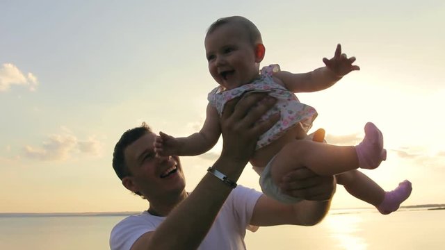 Man lifting his daughter, smiling. Father and baby girl playing on the beach at sunset
