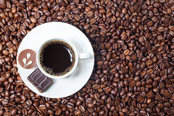 top view of a cup of black coffee and two pieces of chocolate on a white plate placed on coffee bean background,copy space. Clipping path included.