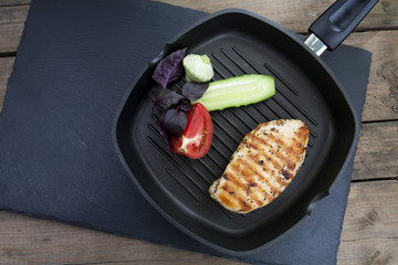 Grilled chicken breast steak with tomato, cucumber and violet basil on teflon pan grill, black slate stone background
