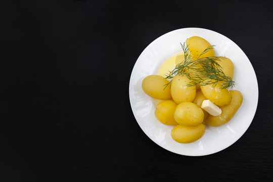 Boiled potatoes with dill and butter in white plate on black slate stone background, top view