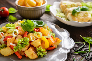 Fried potato salad with lettuce, pepper, onion and baked fish fillets covered with cheese on a wooden background