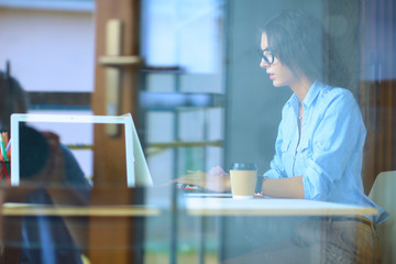 Young woman sitting at office table with laptop, view through window