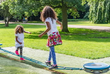 Adorable kids playing outside in park on a summer day. Two brazilian girls with long curly hair wearing a white and colorful dress balancing on a playground, image for family blog - 117121208