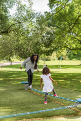Adorable kid playing outside in park on a summer day. Brazilian girl and mother with long curly hair wearing a white dress, blouse and pants balancing on a playground, image for family blog