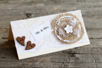 Vintage wedding invitation on old wooden table. Handmade wedding invitation card decorated with lace and burlap flower and wooden hearts. Lovely card crafts. Closeup