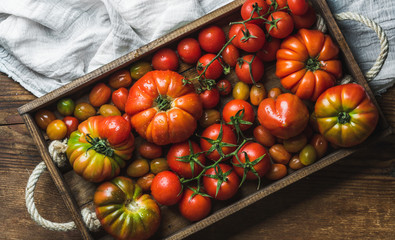 Colorful tomatoes of different sizes and kinds in dark wooden tray over light textile and rustic...