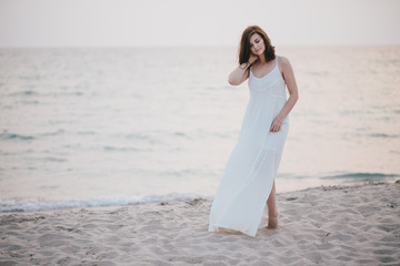 Fototapeta na wymiar Young beautiful woman in a white dress walking on an empty beach near ocean