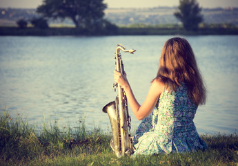 Vintage portrait of a beautiful young woman sitting on the riverbank with a sax