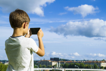 Fototapeta na wymiar Boy with mobile phone. Child taking photo his smartphone. Beautiful sky and city background. Back view. Technology concept