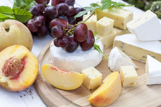 Different delicious cheeses camembert, blue cheese, cheese with nut, fruits red grapes and peach and herbs mint and arugula on wooden round board