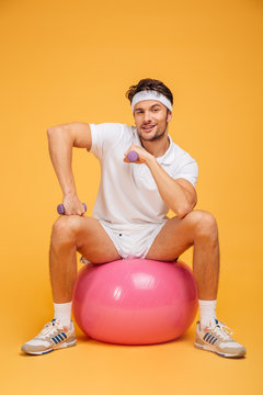 Smiling sportsman sitting on the fitness ball holding dumbbells
