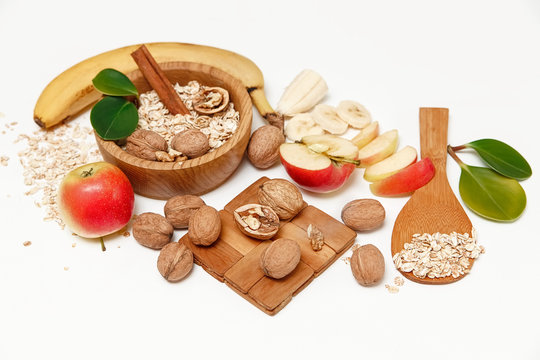 There are Banana,Apple, Walnuts in the Wooden Plate and Rolled Oats,Wooden Spoon,Trivet,with Green Leaves,Healthy Fresh Organic Food on the White Background