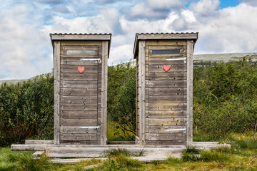 Two wooden outhouse toilets with red heart in mountain landscape. 