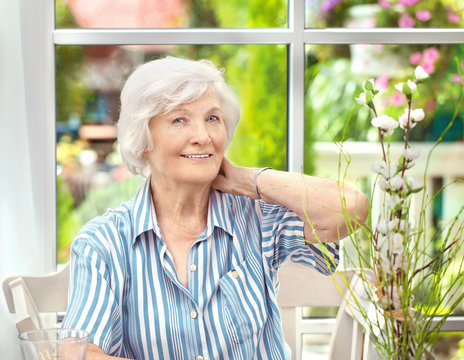  Seniorin sitzt im Wintergarten ,eine Hand im Nacken, im Hintergrund der Garten