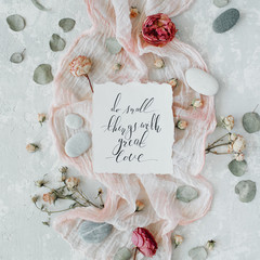 inspirational quote "do small things with great love" written in calligraphy style on paper with dry white tulips, eucalyptus petals and pink textile on concrete background. Flat lay, top view