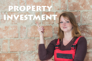 Attractive young redhead woman is standing  in front of an old brick wall. Woman is looking at the camera and is pointing the finger at the sign Property Investment