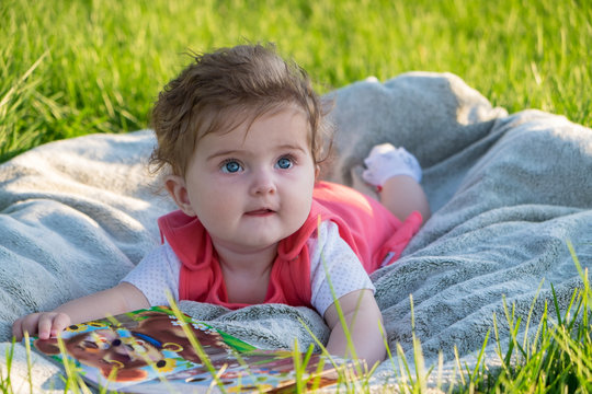 Little girl (six months old) with beautiful blue eyes is looking at pictures in the book. She lies on the grass in the park.