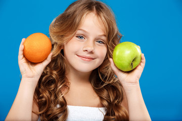 Young pretty girl holding apple and orange over blue background.