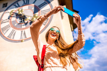 Young female traveler making selfie photo with smart phone on the old clock tower background in...