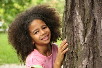 Afro-American little girl hiding behind tree in park
