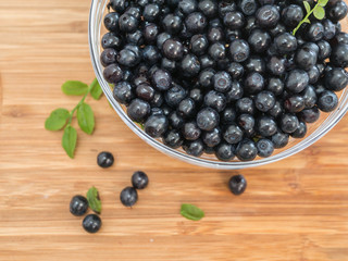 clear glass bowl full of fresh blueberries, branch, green leaves, berries on wooden surface. Close up. View from top. 