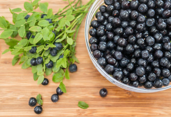 clear glass bowl full of fresh blueberries, green leaves, berries on wooden surface 