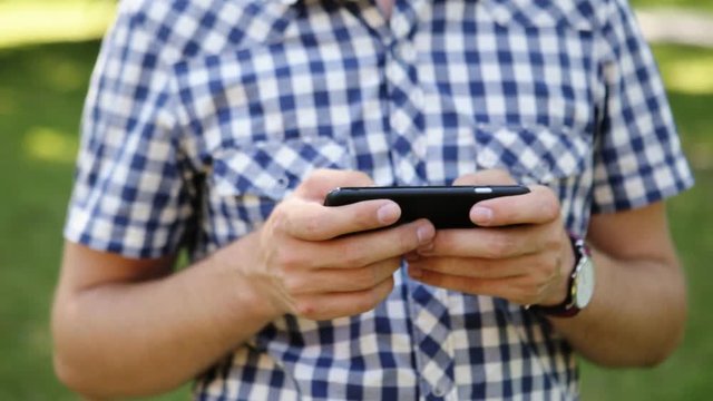Closeup photo of a handsome young man in checkered shirt typing a message, outdoors