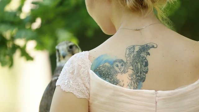 Charming woman in a pink dress with owl tattoo holding a falcon on arm