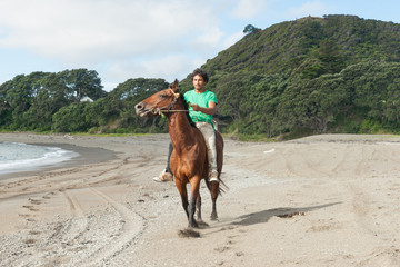 Young Maori man in green tee shirt rides along Ta Kaha beach on brown horse