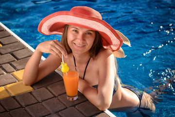 Beautiful young girl in a bathing suit and hat drinking fresh fruit juice from a glass in the luxury  pool