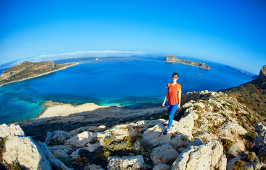 Fototapeta premium sporty female traveler with backpack standing on the cliff against sea and blue sky with white clouds at early morning, Crete, Greece.
