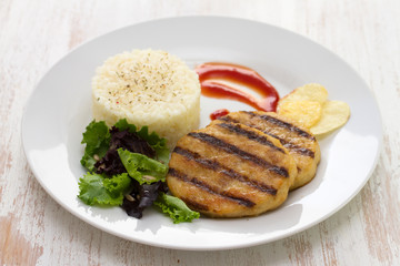 grilled hamburger with rice, salad and chips on plate on white wooden background