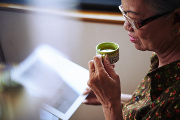 Aged Asian woman drinking tea and using tablet