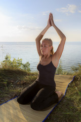 Beautiful young woman practising yoga on mat outdoors at river bank on grass
