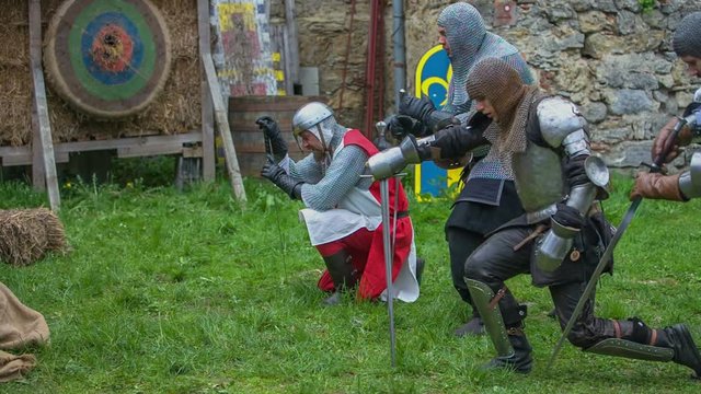 Four men come before the king and they kneel down. Their swords are put into a soil. Close-up shot. A cameraman is in the background.
