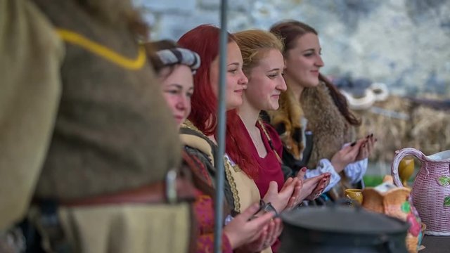 Women of the royal family are sitting at a table in a tent and they are gracefully clapping. The king stands up. Close-up shot. Ladies are also smiling.
