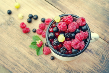 Fresh juicy berries, raspberries, currants, blackberries, a gooseberry in an old white iron mug on a wooden surface