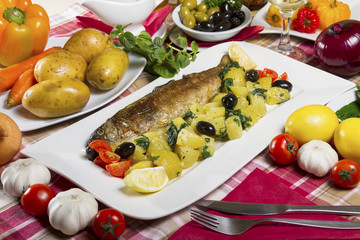 Whole fried trout served on a white plate with potato salad and chard with olives, lemon and cherry tomatoes next to fresh vegetables.