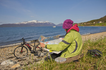 Frau schaut in eine Landschaftskarte. Sie sitzt am Strand. Ein Fahrrad steht vor dem See und Berg