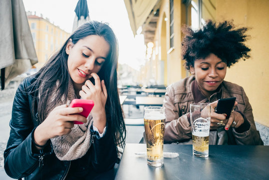 Two multiethnic beautiful young woman black and caucasian sitting in a bar having a beer using smart phone hand hold - technology, social network, communication concept