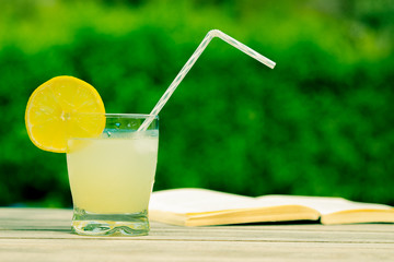 Glass of iced lemonade and opened book on the table outdoors