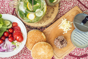 Ingredients for cooking burgers. Raw ground beef meat cutlets on wooden chopping board, red onion, cherry tomatoes, greens, cheese and lemonade on table