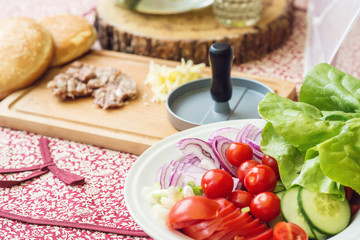 Ingredients for cooking burgers. Raw ground beef meat cutlets on wooden chopping board, red onion, cherry tomatoes, greens, cheese on table