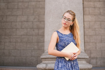 Beautiful and smart girl with glasses keeps book staying near University building. Higher education, college