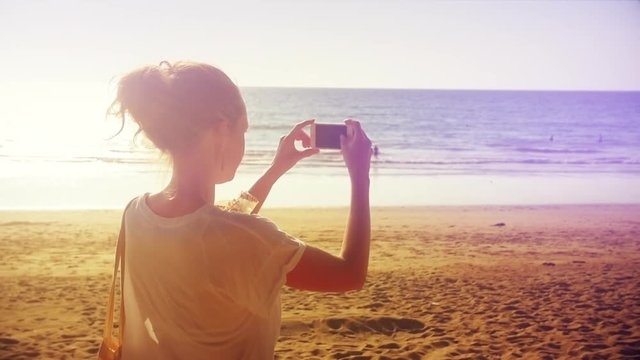 Attractive young caucasian woman is taking pictures with her smartphone on a tropical beach. Vacation, travel lifestyle, or social media concept. Wind in her hair in slow motion. Graded look.